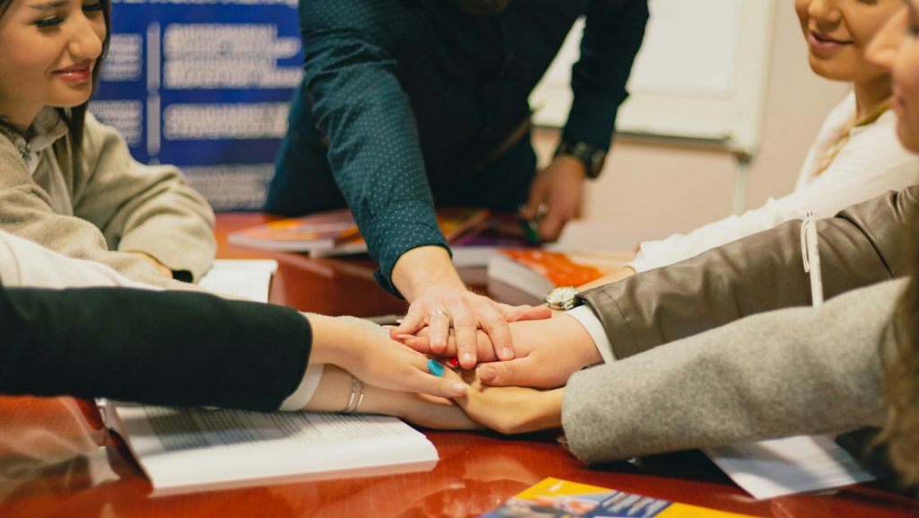 Mitarbeiter, die ihre Hände für den Zusammenhalt in der Mitte des Tisches aufeinander legen.